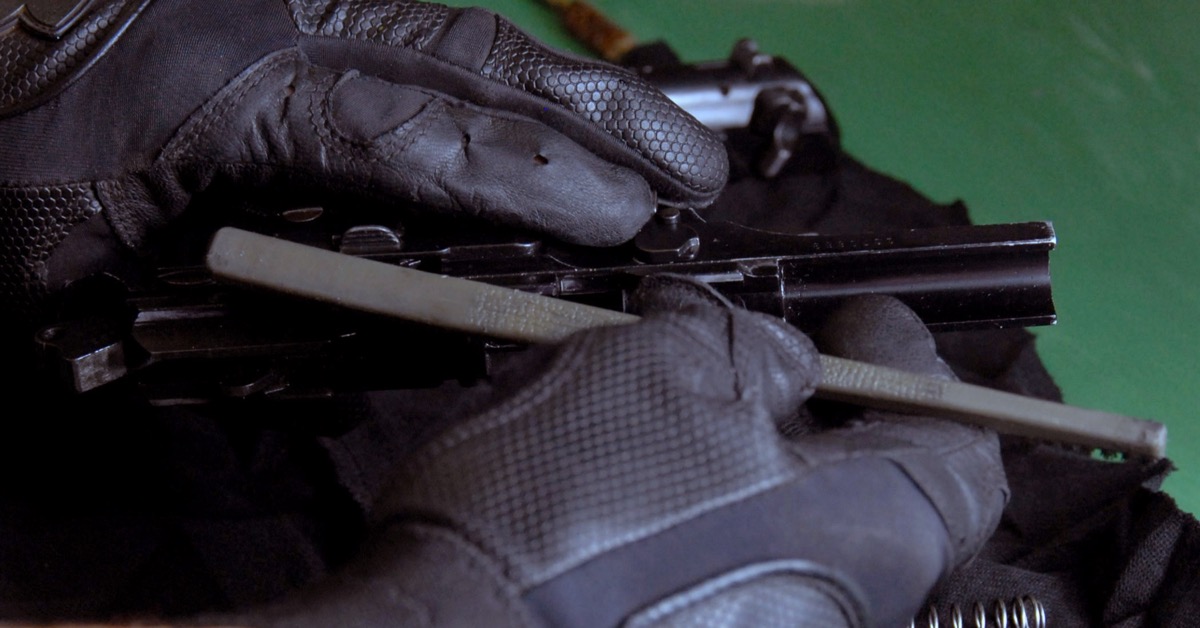 Gloved hands use a brush to clean a disassembled pistol after a range session.